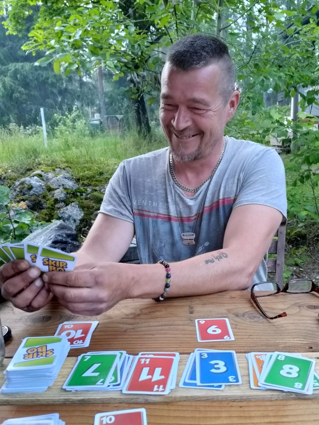 Antti Mikkonen istuu puutarhassa pöydän ääressä korttipeli kädessä – rento ja aito hetki arjen keskellä.